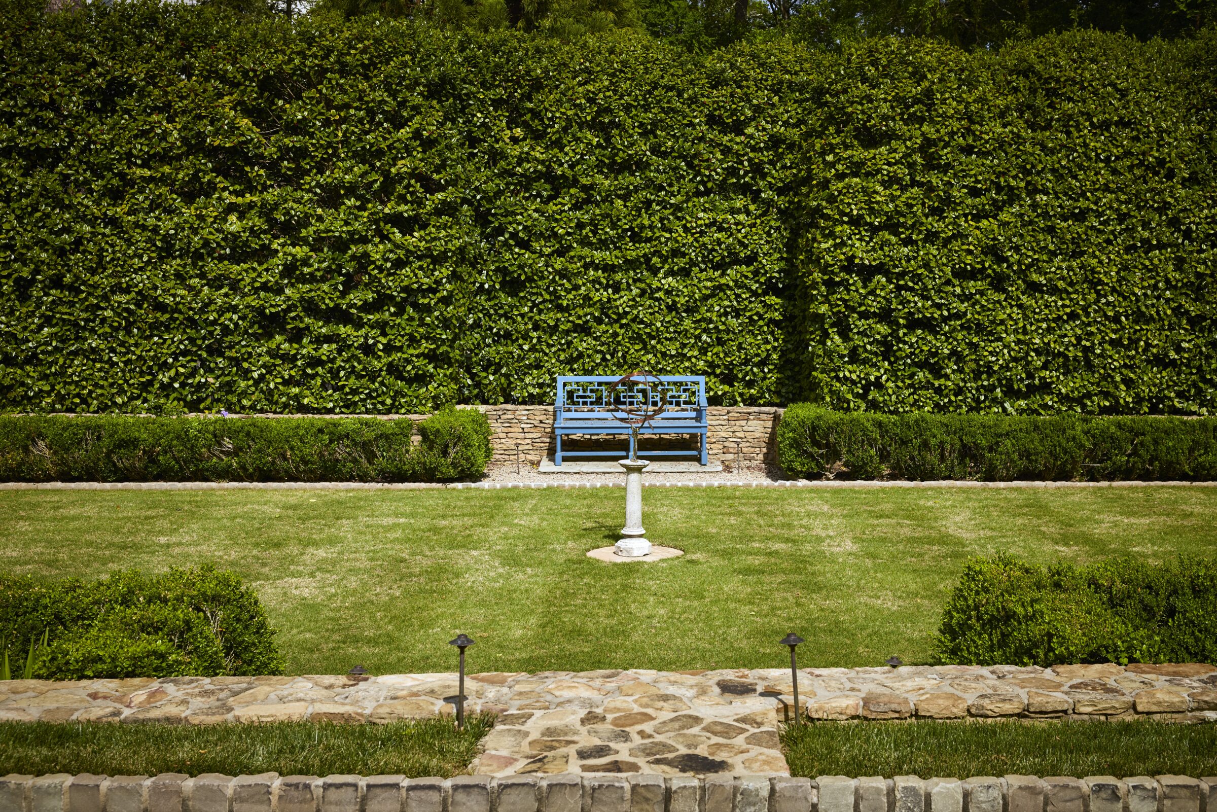 Formal garden landscape with manicured hedges, stone pathways, a blue garden bench, and symmetrical lawn design.