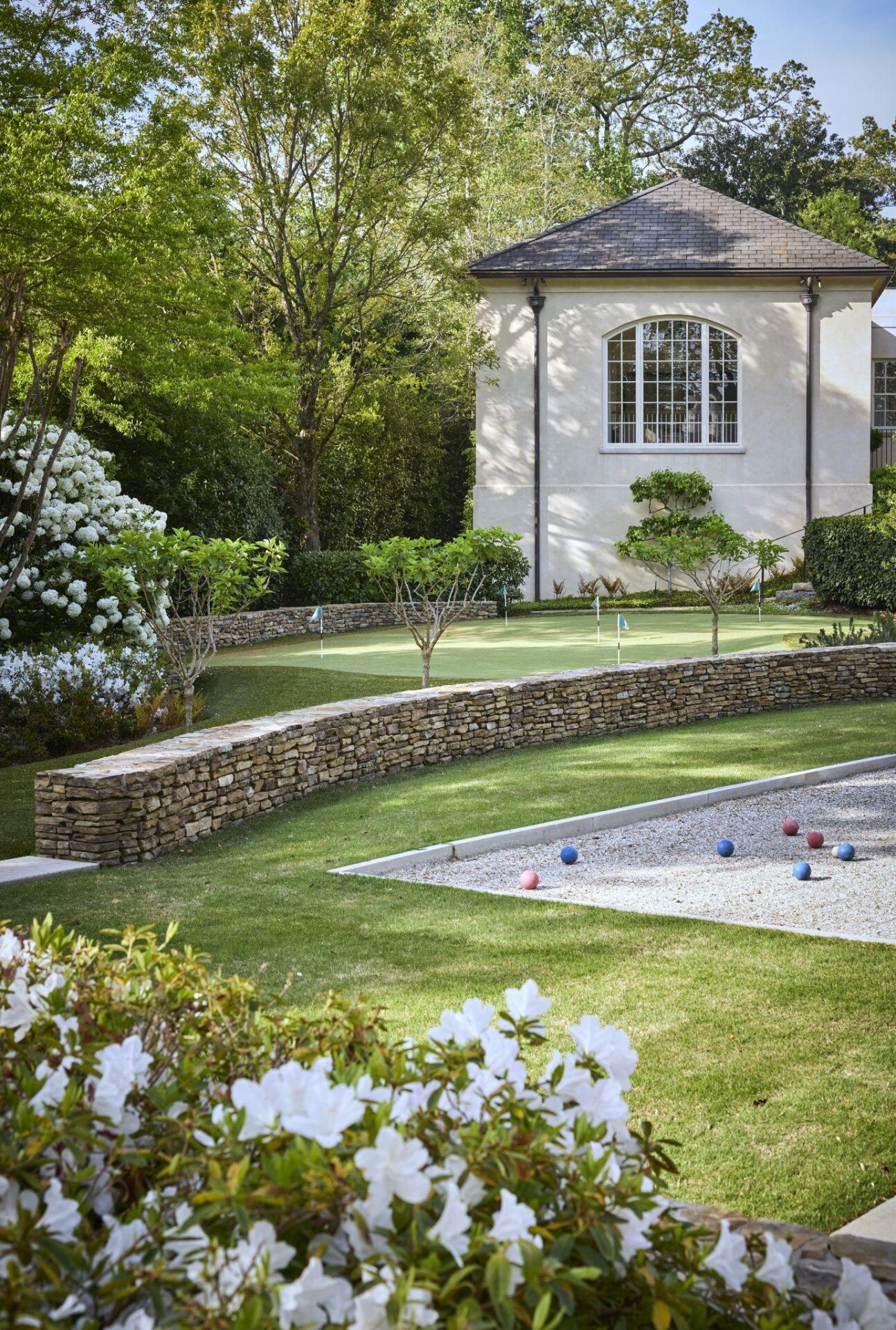 Luxury estate landscape with manicured lawn, bocce court, putting green, stone retaining walls, and blooming white flowers.